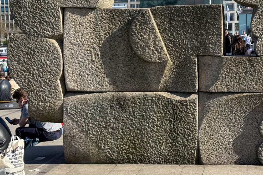 Nahaufnahme einer Skulptur aus großen, unterschiedlich geformten Steinblöcken, die wie ein Puzzle zusammengesetzt sind. Durch Lücken links unten und rechts oben sind Menschen auf einem Platz zu sehen, eine Person benutzt ein Smartphone..